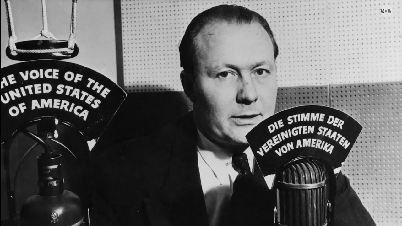Archivo JPG con fotografía del locutor de la VOA con un micrófono que tiene un letrero que dice en idioma alemán: Die Stimme Der Vereinigten staaten von amerika (La Voz de los Estados Unidos de América)